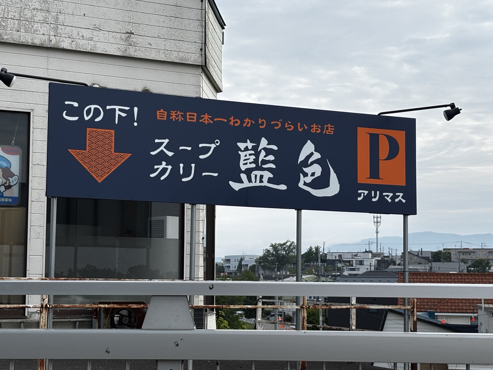 藍色の看板。「この下！自称日本一わかりづらいお店」と書いてある